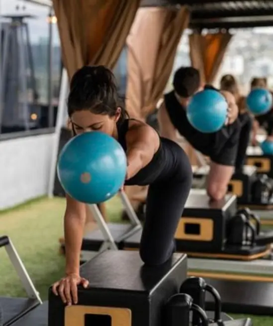 A woman performing a reformer Pilates workout using a blue exercise ball at an outdoor studio, next to a 5-star client review by Brittany B about Natural Pilates in LA.